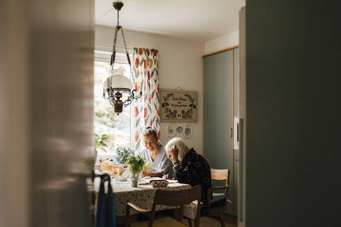 Ältere Frau, die in der Küche zu Hause bei einer weiblichen Betreuerin sitzt und Zeitung liest - MASF27643