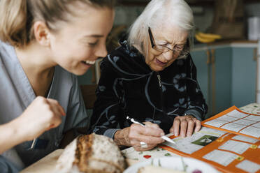 Lächelnde weibliche Betreuerin schaut, während eine ältere Frau in der Küche ein Kreuzworträtsel löst - MASF27649