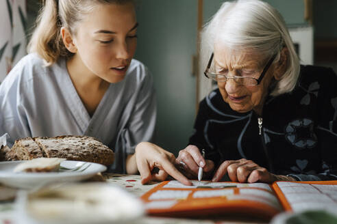 Mitarbeiterin des Gesundheitswesens löst Kreuzworträtsel mit einer älteren Frau, während sie zu Hause in der Küche sitzt - MASF27650
