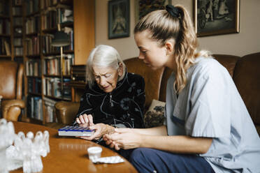 Ältere Frau unterhält sich mit einer weiblichen Betreuerin über einer Hausapotheke im Wohnzimmer - MASF27657