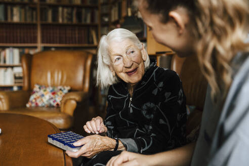 Lächelnde ältere Frau mit Medikamentenschachtel im Gespräch mit einer Mitarbeiterin des Gesundheitswesens im heimischen Wohnzimmer - MASF27658