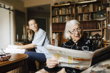 Senior woman reading newspaper while female caregiver looking at her in living room - MASF27664