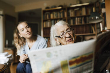 Senior woman reading reading newspaper by female healthcare worker in living room - MASF27666