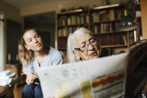 Ältere Frau liest Zeitung, die von einer Mitarbeiterin des Gesundheitswesens im Wohnzimmer gelesen wird - MASF27666