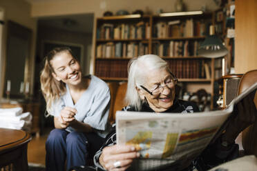 Lächelnde ältere Frau, die Zeitung liest, während im Hintergrund eine glückliche Betreuerin zu Hause ist - MASF27667