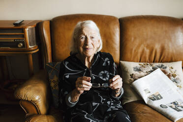Portrait of senior woman holding eyeglasses while sitting on sofa - MASF27673