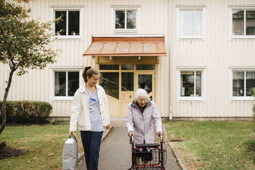 Ältere Frau mit Gehhilfe in Begleitung einer weiblichen Pflegekraft im Vorgarten - MASF27683