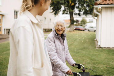 Lächelnde ältere Frau, die eine Krankenschwester ansieht, während sie im Vorgarten spazieren geht - MASF27686