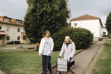 Lächelnde Mitarbeiterin des Gesundheitswesens betrachtet eine ältere Frau mit Rollator auf dem Gehweg - MASF27692