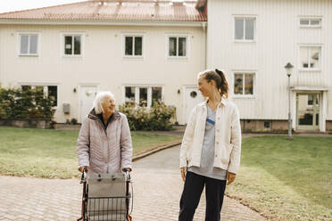 Lächelnde Krankenschwester und ältere Frau schauen sich an, während sie gegen ein Haus laufen - MASF27697