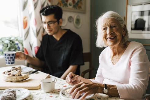 Porträt einer älteren Frau, die mit einem Pfleger in der Küche eines Hauses sitzt - MASF27699