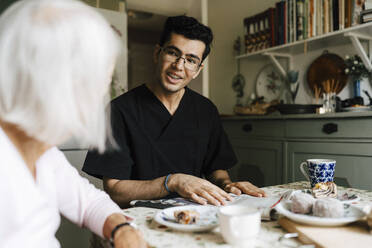 Smiling male caregiver talking with elderly woman in kitchen at home - MASF27702