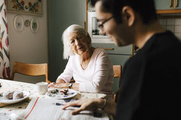 Young male nurse reading newspaper while elderly woman looking at him in kitchen - MASF27704