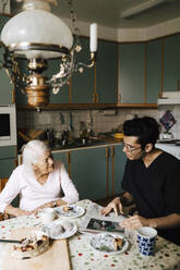 Male healthcare talking with senior woman while reading newspaper at dining table - MASF27705