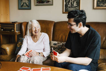 Glückliche männliche Pflegekraft, die mit einer älteren Frau im Wohnzimmer Karten spielt - MASF27708