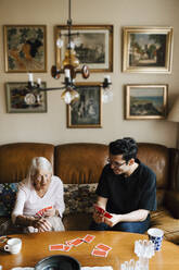 Elderly woman playing cards with male caregiver at table in living room - MASF27710