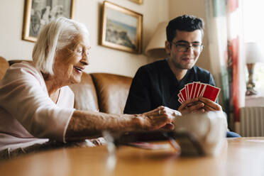 Happy senior woman playing cards with male healthcare worker in living room - MASF27711