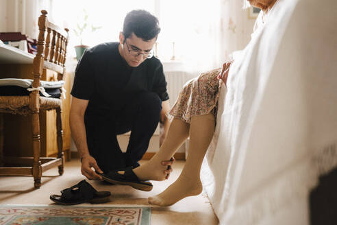 Männlicher Pfleger hilft einer älteren Frau, im Schlafzimmer zu Hause Sandalen zu tragen - MASF27719