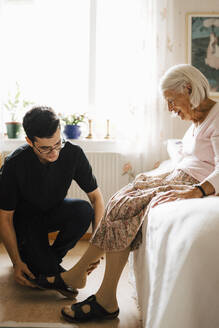 Lächelnde ältere Frau schaut, während männliche Krankenschwester ihr hilft, Sandale im Schlafzimmer zu setzen - MASF27720