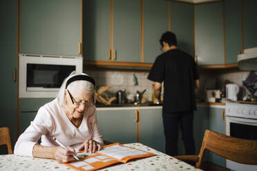 Elderly woman solving crossword puzzle in book while male nurse working at kitchen - MASF27734