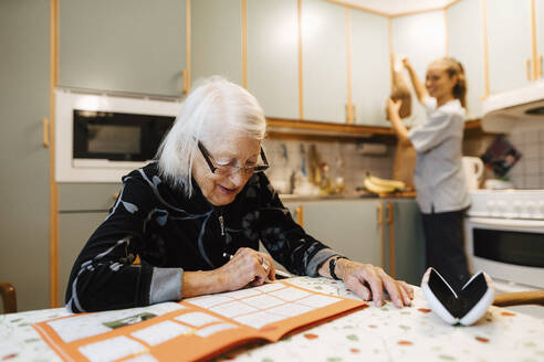 Eine ältere Frau löst ein Sudoku, während eine Krankenschwester in der Küche arbeitet - MASF27740