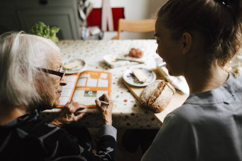 Ältere Frau im Gespräch mit Betreuerin beim Lösen eines Kreuzworträtsels in der Küche - MASF27742