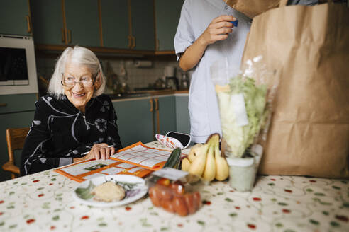 Lächelnde ältere Frau, die wegschaut, sitzt neben einer Krankenschwester in der Küche - MASF27745