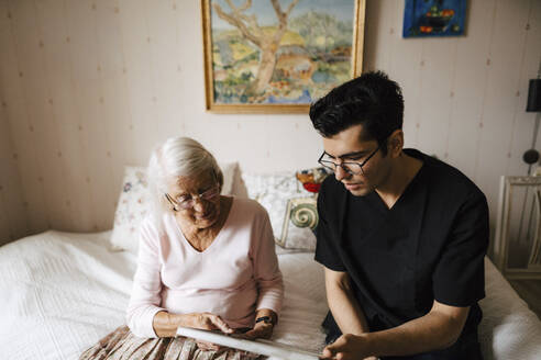 Junger Krankenpfleger zeigt einer älteren Frau, die zu Hause auf dem Bett sitzt, einen Spiegel - MASF27748