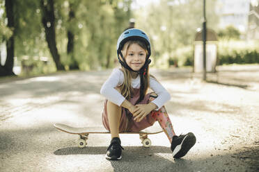 Lächelndes Mädchen mit Beinprothese, das auf einem Skateboard sitzt und nachdenkt - MASF27789