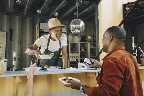 Lächelnder Besitzer, der Essen serviert, während ein männlicher Kunde mit seinem Smartphone kontaktlos bezahlt - MASF27809