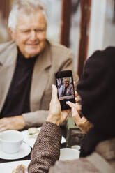 Frau fotografiert älteren Mann mit Smartphone in einem Straßencafé - MASF27841
