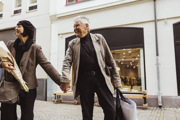 Lächelnder älterer Mann hält die Hände einer Frau beim Gehen auf der Straße in der Stadt - MASF27849