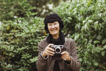 Porträt einer lächelnden älteren Frau mit Kamera vor Pflanzen im Park während des Urlaubs - MASF27866