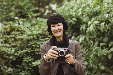 Portrait of smiling senior woman with camera against plants in park during vacation - MASF27866