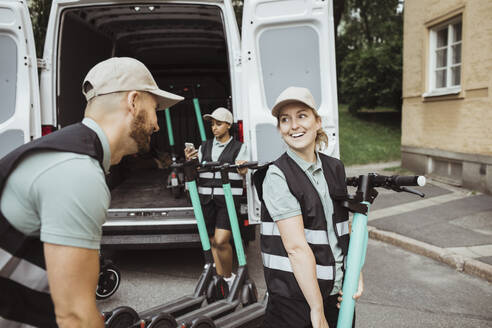 Lächelnde Zustellerin, die einen männlichen Mitarbeiter anschaut, während sie einen Motorroller in einen Lieferwagen lädt - MASF27901