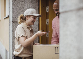 Eine lächelnde Mitarbeiterin fotografiert ein Paket bei der Auslieferung an einen männlichen Kunden - MASF27949