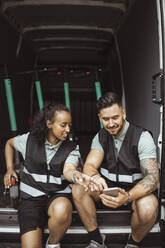 Lächelnde weibliche und männliche Mitarbeiter, die ihr Smartphone benutzen, während sie im Lieferwagen sitzen - MASF27967