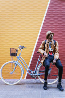 Full body of calm African American male in hat looking into distance while sitting on bicycle on sidewalk near colorful wall - ADSF32580