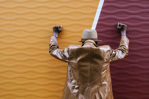Rückenansicht eines anonymen Mannes in stilvollem Ledermantel und Hut, der seine Hände an eine bunte Wand lehnt, während er auf der Straße steht - ADSF32584