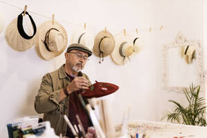 Konzentrierte reife männliche Künstlerin in legerer Kleidung und mit Brille malt auf stilvolle Baskenmütze mit Pinsel während der Arbeit im Kreativstudio - ADSF32586