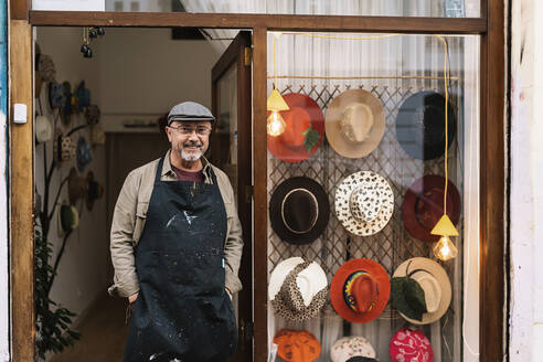 Content mature bearded male master in apron standing near showcase with collection of stylish handmade hats and looking at camera - ADSF32587