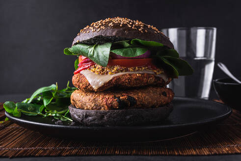 Appetitliche Burger mit Sesam auf schwarzem Teller in der Nähe von Salatblättern gegen Glas Wasser auf dunklem Hintergrund platziert - ADSF32596
