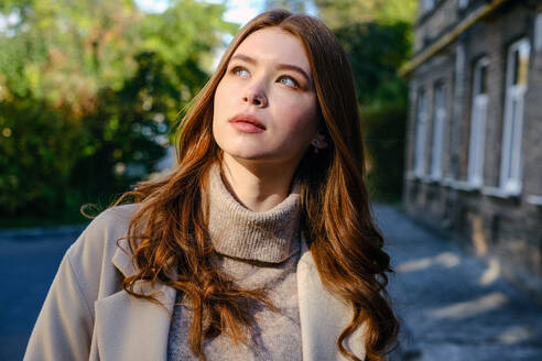 Attractive female with red hair in trendy coat looking up while standing on street near shabby house and green trees - ADSF32616