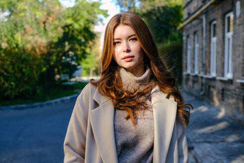 Attractive female with red hair in trendy coat looking at camera while standing on street near shabby house and green trees - ADSF32617