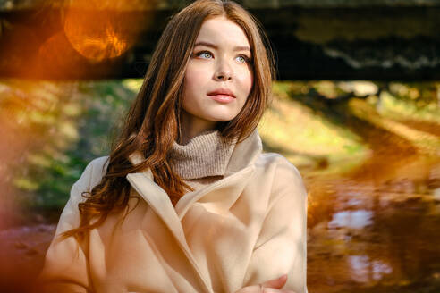 Dreamy female with long red hair in stylish warm coat looking away while standing on street against blurred background with lens flare - ADSF32619