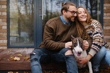 Content couple looking away while sitting on stairs with Bull Terrier near house glass door in fall - ADSF32621