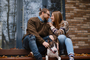 Zufriedenes Paar, das sich auf der Treppe sitzend mit einem Bull Terrier in der Nähe der Glastür eines Hauses im Herbst ansieht - ADSF32622
