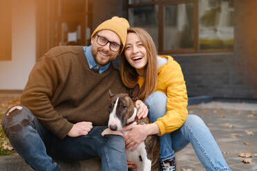 Zufriedenes Paar, das in die Kamera schaut, auf einer Treppe sitzend mit einem Bull Terrier in der Nähe der Glastür eines Hauses im Herbst - ADSF32623