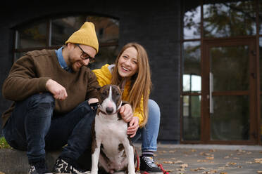 Zufriedenes Paar, das in die Kamera schaut, auf einer Treppe sitzend mit einem Bull Terrier in der Nähe der Glastür eines Hauses im Herbst - ADSF32624