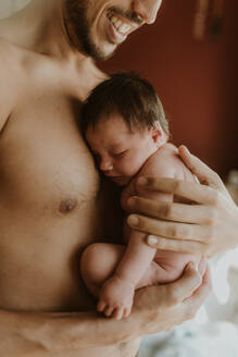 Side view of cropped unrecognizable calm ethnic father with naked torso embracing adorable newborn in bedroom - ADSF32634